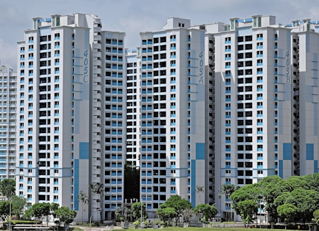High-rise residential buildings with a modern design featuring a combination of white and light blue colors. The buildings are situated in an urban area with lush green trees in the foreground.