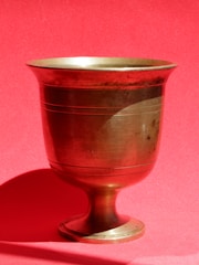 A brass chalice with a simple, elegant design rests against a vibrant red background. The chalice displays a smooth surface with subtle horizontal grooves accentuating its structure. The base is broad and sturdy, supporting the cup's tall, narrow form. The warm metallic sheen of the brass contrasts sharply with the matte red backdrop, creating a striking visual effect.