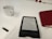 A white table features a white ceramic cup, a digital e-reader displaying text, and a red closed notebook. A power outlet is visible in the background, and a white rectangular object is partially shown in the foreground.