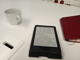 A white table features a white ceramic cup, a digital e-reader displaying text, and a red closed notebook. A power outlet is visible in the background, and a white rectangular object is partially shown in the foreground.