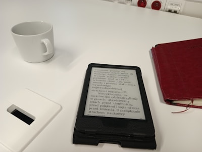 A white table features a white ceramic cup, a digital e-reader displaying text, and a red closed notebook. A power outlet is visible in the background, and a white rectangular object is partially shown in the foreground.