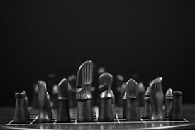 A set of dark, minimalist chess pieces is arranged on a board against a black background. The chess pieces have unique, abstract designs and are made from a smooth material, possibly wood or stone.