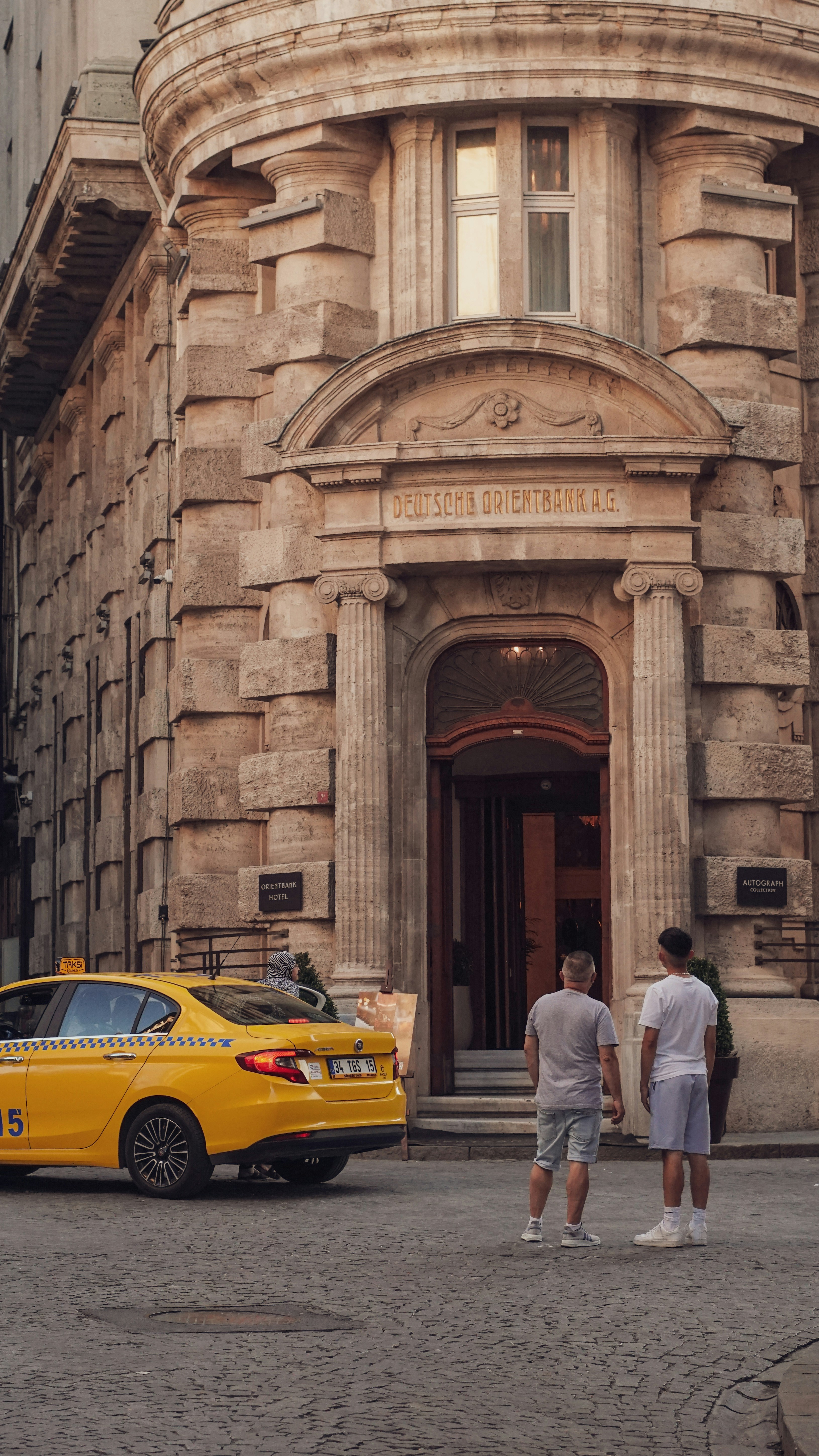 A yellow taxi is parked in front of an old, historical building with a stone facade. Two men are standing on the cobblestone street near the entrance, which has an arched doorway with the inscription 'Deutsche Orientbank A.G.' above it. The building features classic architectural details with large columns and arched windows.
