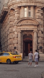 A yellow taxi is parked in front of an old, historical building with a stone facade. Two men are standing on the cobblestone street near the entrance, which has an arched doorway with the inscription 'Deutsche Orientbank A.G.' above it. The building features classic architectural details with large columns and arched windows.