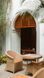 An outdoor seating area features rattan chairs and a round table set against a white stucco wall. An arched doorway with wooden slats provides a shaded entrance, while surrounding green foliage adds a touch of nature.