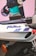 A sleek motorcycle featuring the word 'FireBlade' in a stylized font is visible. The body is predominantly white with blue, red, and purple accents. The tire and a turn signal light add an element of depth. In the background, there's a display of clothing and accessories, including a helmet and folded garments. The setting appears to be an indoor showroom with a promotional display.