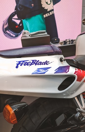A sleek motorcycle featuring the word 'FireBlade' in a stylized font is visible. The body is predominantly white with blue, red, and purple accents. The tire and a turn signal light add an element of depth. In the background, there's a display of clothing and accessories, including a helmet and folded garments. The setting appears to be an indoor showroom with a promotional display.