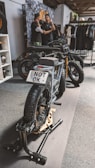 A stylish black electric motorcycle is positioned on a display stand within an indoor setting. The license plate reads 'NOT OK'. In the background, two individuals are engaged in conversation next to a wall adorned with various images and a rack displaying clothing items.