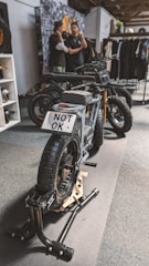 A stylish black electric motorcycle is positioned on a display stand within an indoor setting. The license plate reads 'NOT OK'. In the background, two individuals are engaged in conversation next to a wall adorned with various images and a rack displaying clothing items.