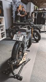 A stylish black electric motorcycle is positioned on a display stand within an indoor setting. The license plate reads 'NOT OK'. In the background, two individuals are engaged in conversation next to a wall adorned with various images and a rack displaying clothing items.
