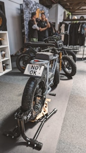 A stylish black electric motorcycle is positioned on a display stand within an indoor setting. The license plate reads 'NOT OK'. In the background, two individuals are engaged in conversation next to a wall adorned with various images and a rack displaying clothing items.