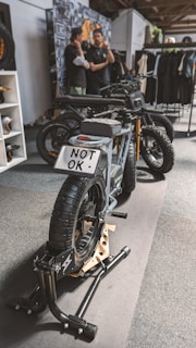A stylish black electric motorcycle is positioned on a display stand within an indoor setting. The license plate reads 'NOT OK'. In the background, two individuals are engaged in conversation next to a wall adorned with various images and a rack displaying clothing items.