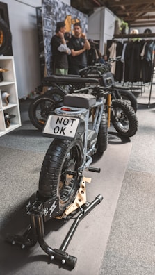 A stylish black electric motorcycle is positioned on a display stand within an indoor setting. The license plate reads 'NOT OK'. In the background, two individuals are engaged in conversation next to a wall adorned with various images and a rack displaying clothing items.