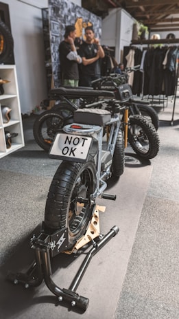 A stylish black electric motorcycle is positioned on a display stand within an indoor setting. The license plate reads 'NOT OK'. In the background, two individuals are engaged in conversation next to a wall adorned with various images and a rack displaying clothing items.