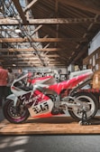 A brightly colored motorcycle is displayed on a wooden platform inside a spacious building with a rustic wooden ceiling. The motorcycle features a striking red, white, and purple design with various decals and the number 347 prominently visible. In the background, people are gathered, and signage for 'Bike Shed Moto Shop' can be seen along with various posters on the walls.