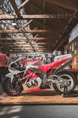 A brightly colored motorcycle is displayed on a wooden platform inside a spacious building with a rustic wooden ceiling. The motorcycle features a striking red, white, and purple design with various decals and the number 347 prominently visible. In the background, people are gathered, and signage for 'Bike Shed Moto Shop' can be seen along with various posters on the walls.
