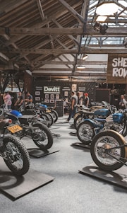 A motorcycle exhibition features several vintage-style bikes displayed inside a wooden-roofed hall. People are casually walking around, observing the motorcycles. The setting includes banners and signage for Devitt Insurance. The atmosphere is well-lit, highlighting the classic design and detailing of the bikes.
