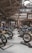 A motorcycle exhibition features several vintage-style bikes displayed inside a wooden-roofed hall. People are casually walking around, observing the motorcycles. The setting includes banners and signage for Devitt Insurance. The atmosphere is well-lit, highlighting the classic design and detailing of the bikes.