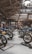A motorcycle exhibition features several vintage-style bikes displayed inside a wooden-roofed hall. People are casually walking around, observing the motorcycles. The setting includes banners and signage for Devitt Insurance. The atmosphere is well-lit, highlighting the classic design and detailing of the bikes.