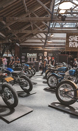 A motorcycle exhibition features several vintage-style bikes displayed inside a wooden-roofed hall. People are casually walking around, observing the motorcycles. The setting includes banners and signage for Devitt Insurance. The atmosphere is well-lit, highlighting the classic design and detailing of the bikes.