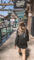 A person with long hair walks down a stone-paved pathway, wearing a black jacket and carrying a black backpack. The setting appears to be an upscale marketplace or club area with a sign overhead reading 'The Bike Shed Motorcycle Club'. The area is bustling with people and has a mix of industrial and rustic architectural elements including exposed beams, brick walls, and ambient lighting.