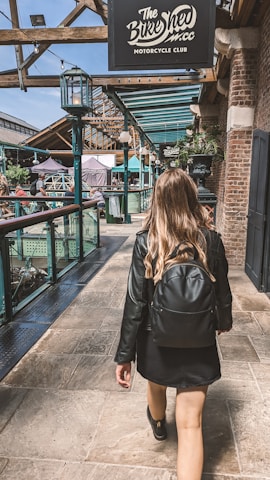 A person with long hair walks down a stone-paved pathway, wearing a black jacket and carrying a black backpack. The setting appears to be an upscale marketplace or club area with a sign overhead reading 'The Bike Shed Motorcycle Club'. The area is bustling with people and has a mix of industrial and rustic architectural elements including exposed beams, brick walls, and ambient lighting.