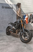 A sleek, black Yamaha motorcycle is positioned on a concrete floor near a textured wall. The bike features prominent golden front forks and a minimalist design. Behind it hangs a collection of jackets and shirts on a clothing rack.
