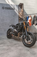 A sleek, black Yamaha motorcycle is positioned on a concrete floor near a textured wall. The bike features prominent golden front forks and a minimalist design. Behind it hangs a collection of jackets and shirts on a clothing rack.