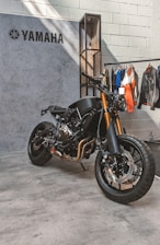 A sleek, black Yamaha motorcycle is positioned on a concrete floor near a textured wall. The bike features prominent golden front forks and a minimalist design. Behind it hangs a collection of jackets and shirts on a clothing rack.