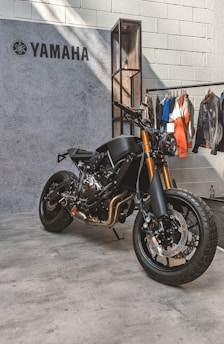 A sleek, black Yamaha motorcycle is positioned on a concrete floor near a textured wall. The bike features prominent golden front forks and a minimalist design. Behind it hangs a collection of jackets and shirts on a clothing rack.
