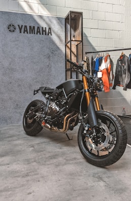 A sleek, black Yamaha motorcycle is positioned on a concrete floor near a textured wall. The bike features prominent golden front forks and a minimalist design. Behind it hangs a collection of jackets and shirts on a clothing rack.