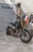 A sleek, black Yamaha motorcycle is positioned on a concrete floor near a textured wall. The bike features prominent golden front forks and a minimalist design. Behind it hangs a collection of jackets and shirts on a clothing rack.