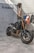 A sleek, black Yamaha motorcycle is positioned on a concrete floor near a textured wall. The bike features prominent golden front forks and a minimalist design. Behind it hangs a collection of jackets and shirts on a clothing rack.