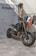 A sleek, black Yamaha motorcycle is positioned on a concrete floor near a textured wall. The bike features prominent golden front forks and a minimalist design. Behind it hangs a collection of jackets and shirts on a clothing rack.