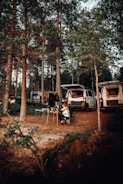 A forest setting with several vans parked among tall trees. A person sits at a table with camping equipment and supplies in front of one of the vans, suggesting a camping scene.