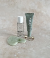 A collection of skincare products displayed on a light speckled surface. The items include a small bottle of micellar water, a tube labeled 'PEEL' for a brightening AHA peel mask, a green jade gua sha tool, and a gold massage tool with two spherical ends.