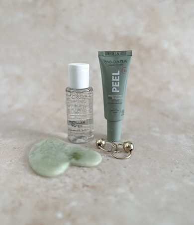 A collection of skincare products displayed on a light speckled surface. The items include a small bottle of micellar water, a tube labeled 'PEEL' for a brightening AHA peel mask, a green jade gua sha tool, and a gold massage tool with two spherical ends.