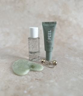 A collection of skincare products displayed on a light speckled surface. The items include a small bottle of micellar water, a tube labeled 'PEEL' for a brightening AHA peel mask, a green jade gua sha tool, and a gold massage tool with two spherical ends.