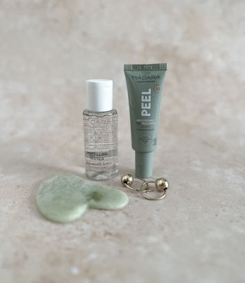 A collection of skincare products displayed on a light speckled surface. The items include a small bottle of micellar water, a tube labeled 'PEEL' for a brightening AHA peel mask, a green jade gua sha tool, and a gold massage tool with two spherical ends.