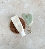 A flat lay composition featuring a tube of cream or lotion on a woven mat, a jade gua sha tool in a heart shape, and a pair of gold hoop earrings with spherical accents, all placed on a textured, light-colored stone surface.