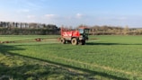 A large red agricultural vehicle is operating in a vast green field, likely performing tasks such as spraying or spreading. The field is surrounded by trees under a clear blue sky with some scattered clouds.