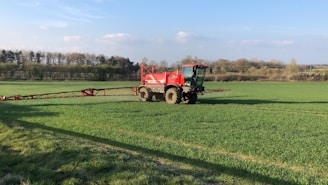 A large red agricultural vehicle is operating in a vast green field, likely performing tasks such as spraying or spreading. The field is surrounded by trees under a clear blue sky with some scattered clouds.