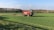 A large red agricultural vehicle is operating in a vast green field, likely performing tasks such as spraying or spreading. The field is surrounded by trees under a clear blue sky with some scattered clouds.