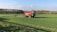 A large red agricultural vehicle is operating in a vast green field, likely performing tasks such as spraying or spreading. The field is surrounded by trees under a clear blue sky with some scattered clouds.