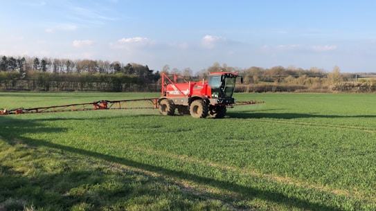 A large red agricultural vehicle is operating in a vast green field, likely performing tasks such as spraying or spreading. The field is surrounded by trees under a clear blue sky with some scattered clouds.