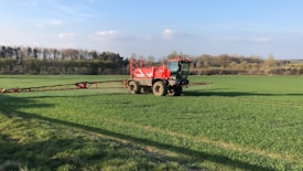 A large red agricultural vehicle is operating in a vast green field, likely performing tasks such as spraying or spreading. The field is surrounded by trees under a clear blue sky with some scattered clouds.
