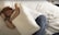 A person with short, red hair sleeps on a white, textured bed while hugging a white pillow. They wear a white sleeveless top, blue jeans, and a black wristband. The setting appears calm and serene, with a soft lighting casting shadows on the sheets.