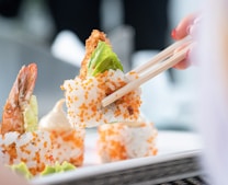 A pair of chopsticks is holding a piece of sushi, garnished with a crispy shrimp tail, lettuce, and orange fish roe. The sushi roll is neatly coated with rice and sesame seeds. The background is slightly blurred, focusing on the appetizing sushi piece.