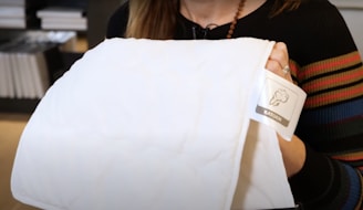 A person is holding a white cotton fabric with a tag labeled 'Katoen.' The person is wearing a multi-colored striped sweater, and there are shelves with books or boxes in the background.