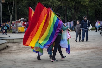 A vibrant scene in a park with a group of people holding large, colorful rainbow flags. In the background, other people are visible walking and chatting. Trees provide a lush backdrop, and there are vendors with carts selling refreshments. The atmosphere is lively and inclusive.